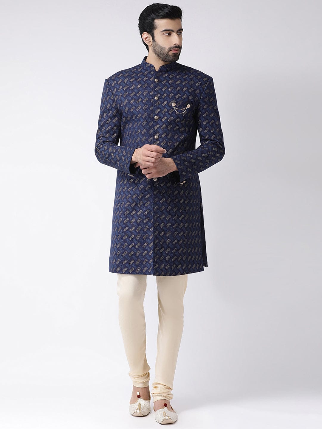 A man modeling a royal blue sherwani with a gold geometric pattern, paired with cream trousers and white shoes, against a light gray studio background.