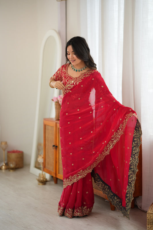 A woman wearing a bright red traditional sari with gold embroidery and a black border, standing in a softly lit indoor room with curtains and a wooden cabinet in the background.