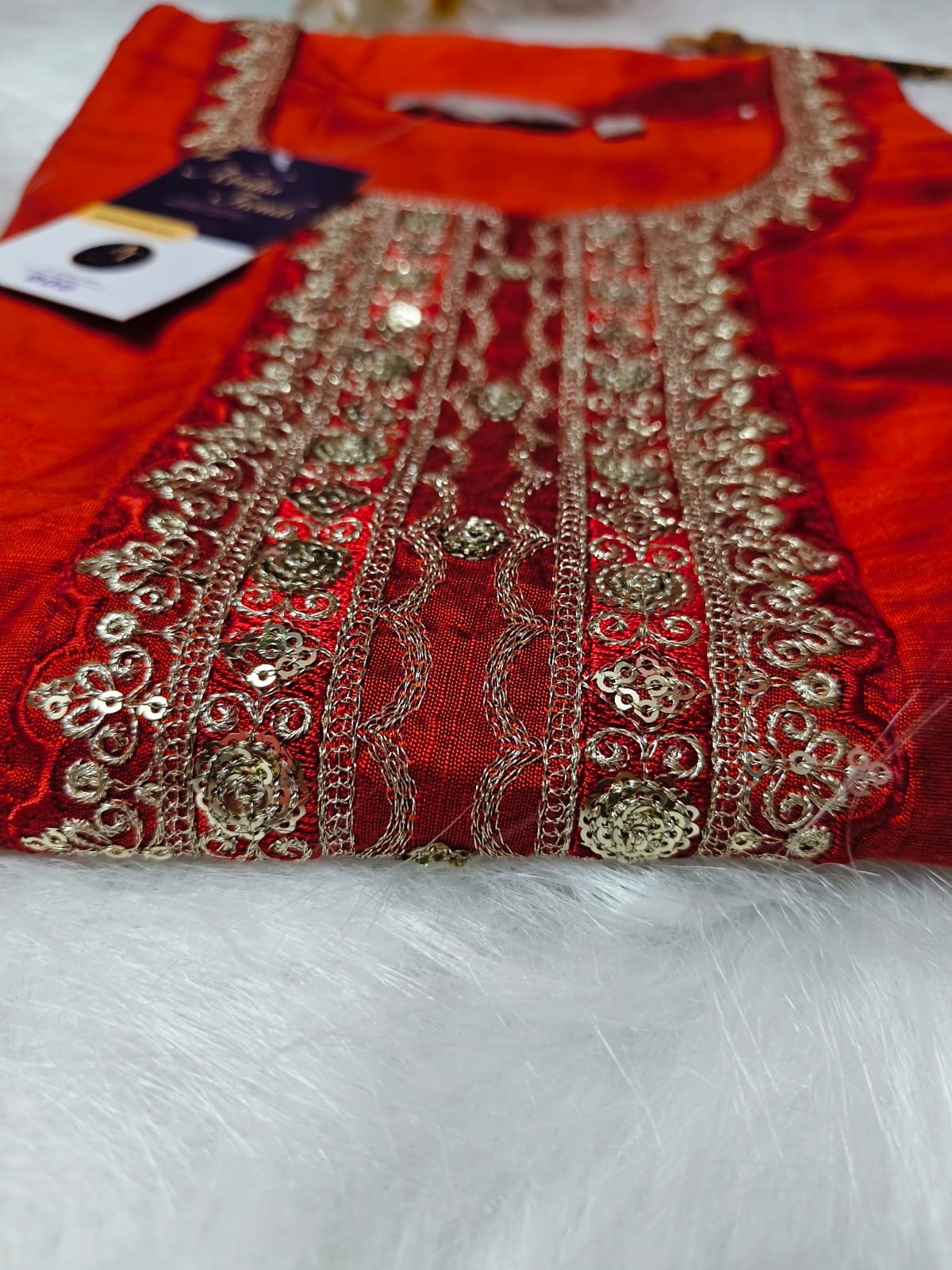 A folded red traditional embroidered garment, likely a kurta or suit piece, featuring ornate gold metallic embroidery and sequins on the front panel, with a dark brand tag attached. The garment is laid flat on a white faux fur surface.