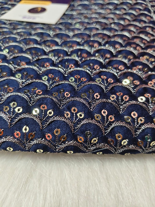 A close-up of a navy blue fabric with an embroidered scallop or fan pattern in metallic silver, decorated with small sequins in gold, pink, and green, laid flat on a white faux fur surface.