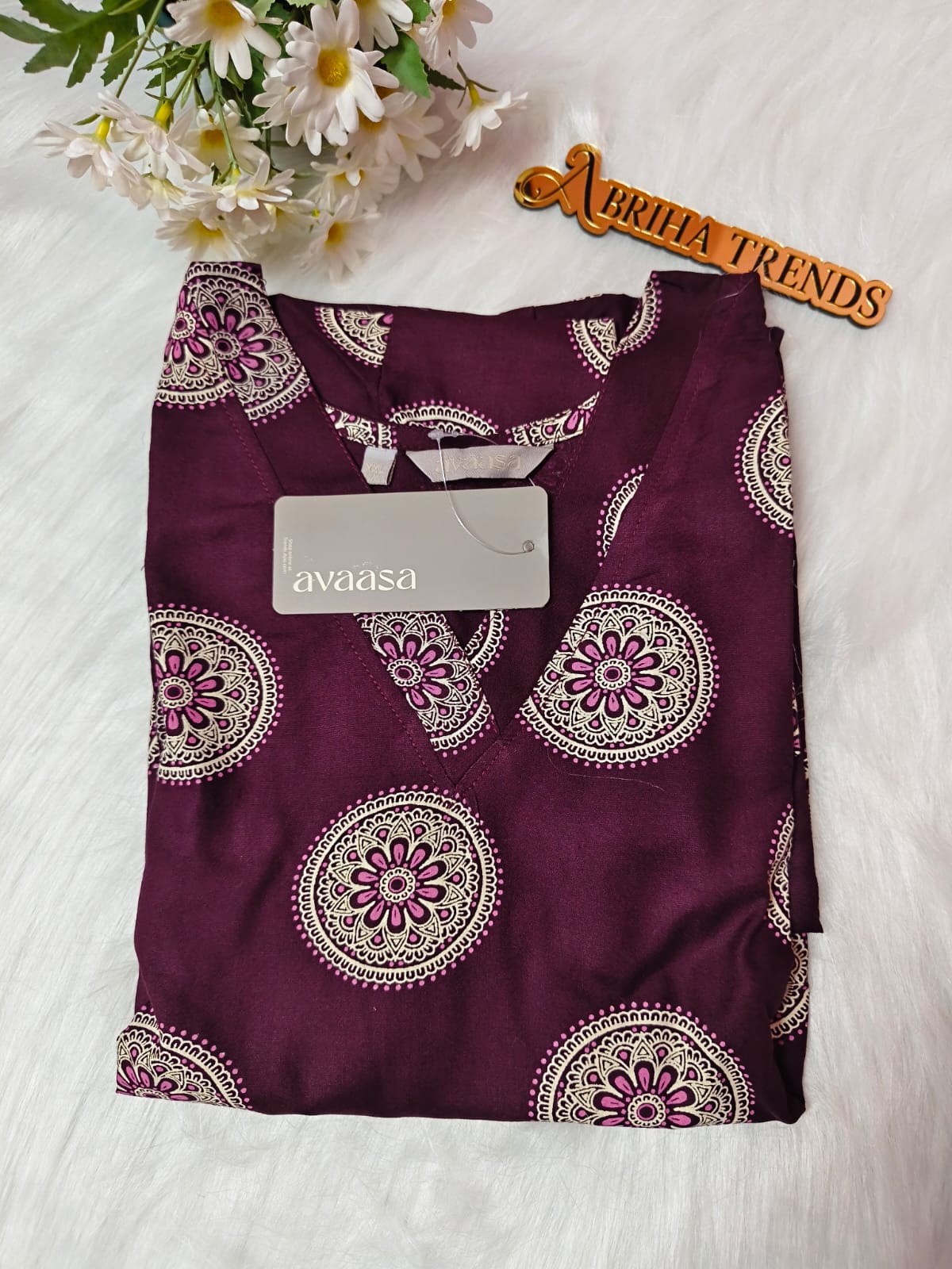 A folded dark purple kurta with a white and pink mandala pattern, featuring a white 'avaasa' brand tag, laid flat on a white faux fur surface with daisies and a 'Abriha Trends' wooden sign in the background.