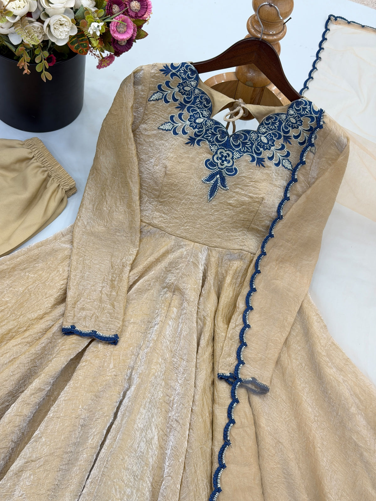 A beige long-sleeve dress with dark blue floral embroidery around the neckline and blue scalloped trim down the front, displayed on a wooden hanger with a matching beige sash or belt partially visible.