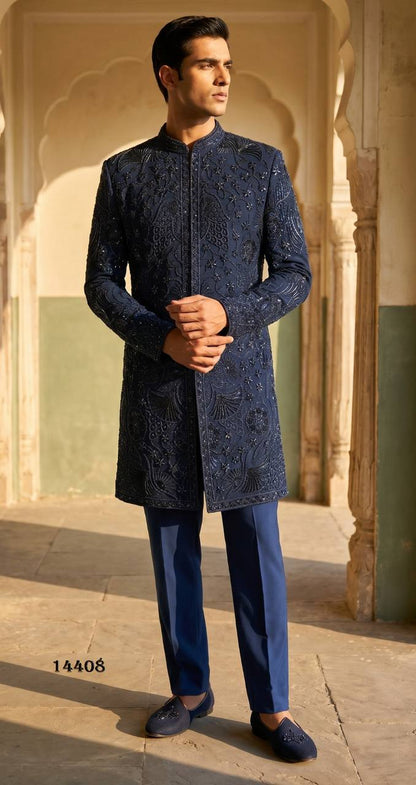 A man modeling a royal blue embroidered sherwani jacket with matching trousers and royal loafers, standing in a sunlit courtyard with arches.