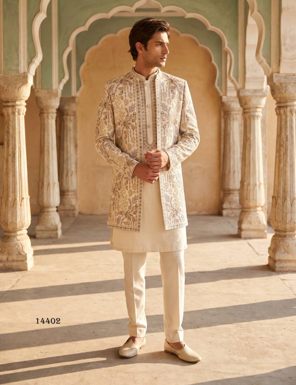 A man wearing a cream and beige heavily embroidered sherwani jacket with matching cream kurta and trousers, standing in a historic courtyard with arches.