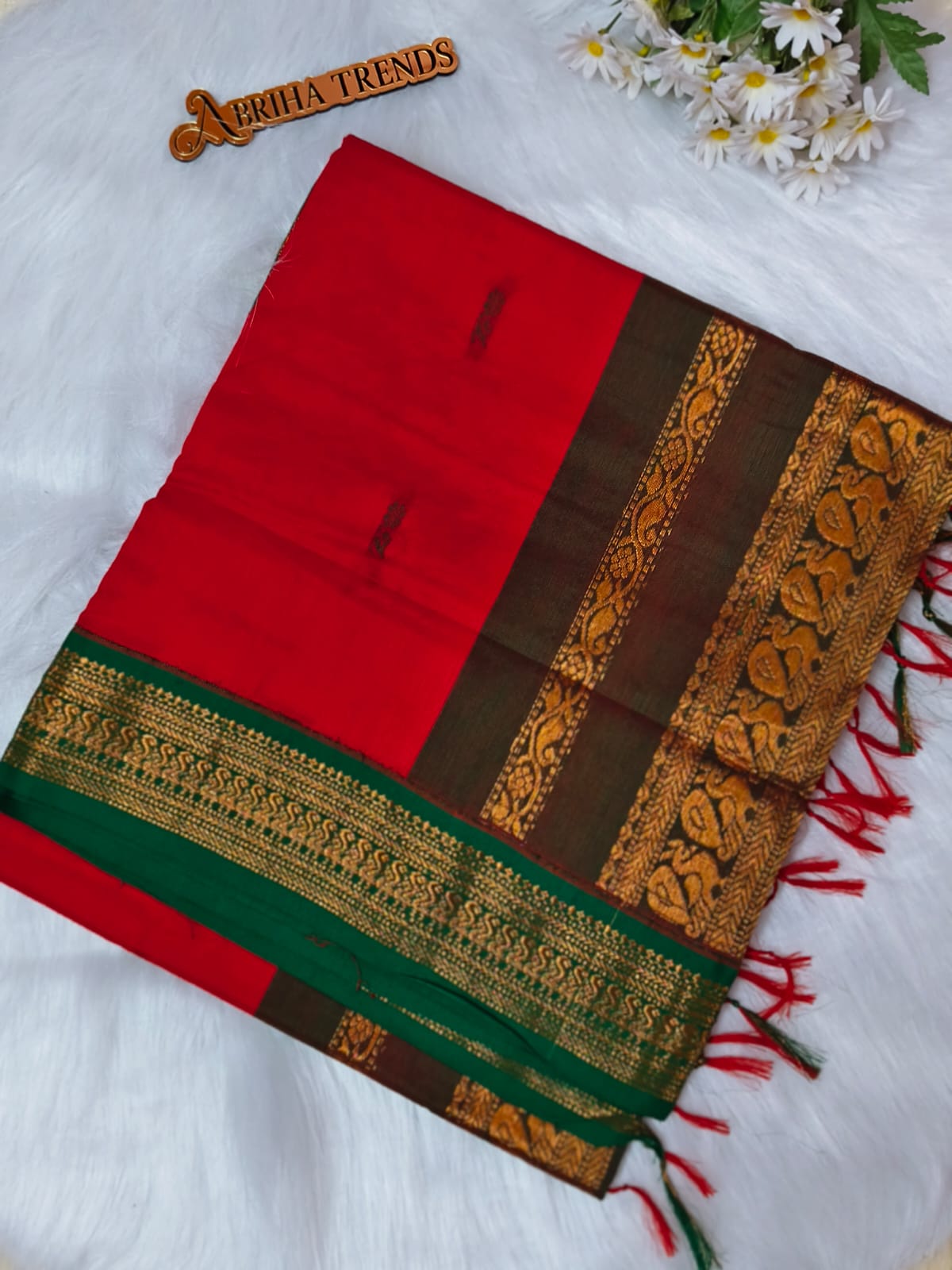 A folded red and brown silk saree with a green border and intricate gold zari embroidery, displayed on a white faux fur surface with a decorative wooden tag and white daisies.