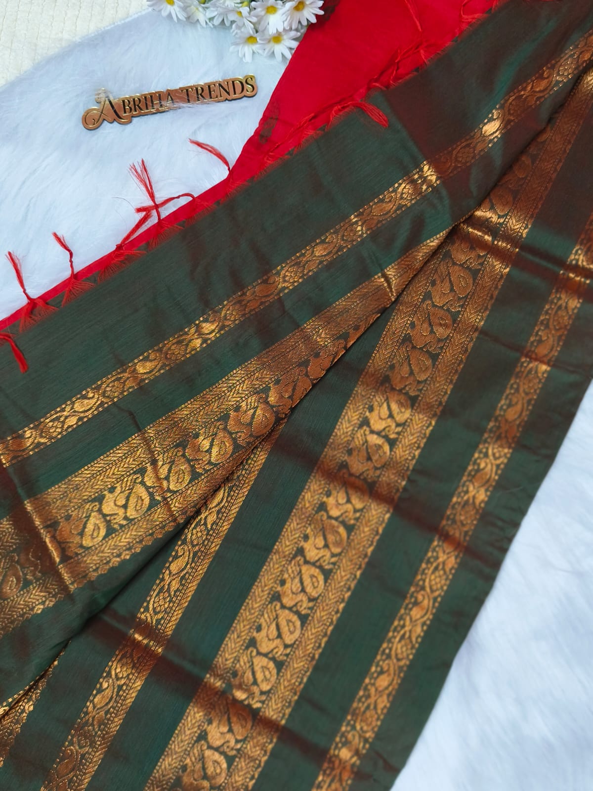A dark green and red saree with intricate gold zari woven borders, draped on a white textured surface with a small decorative wooden tag reading 'BRIEF TRENDS'.