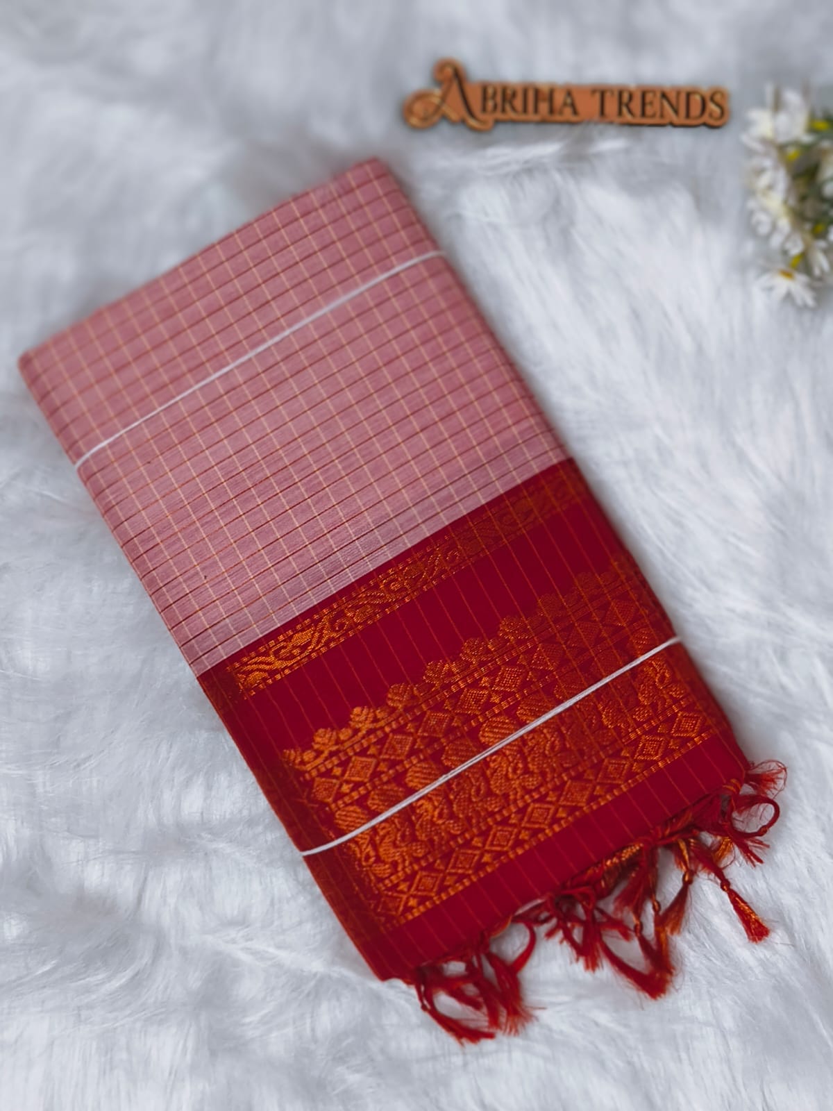 A folded red and pink saree with a gold floral border and white accent stripes, displayed on a white faux fur surface with a 'ABRIHA TRENDS' wooden sign.