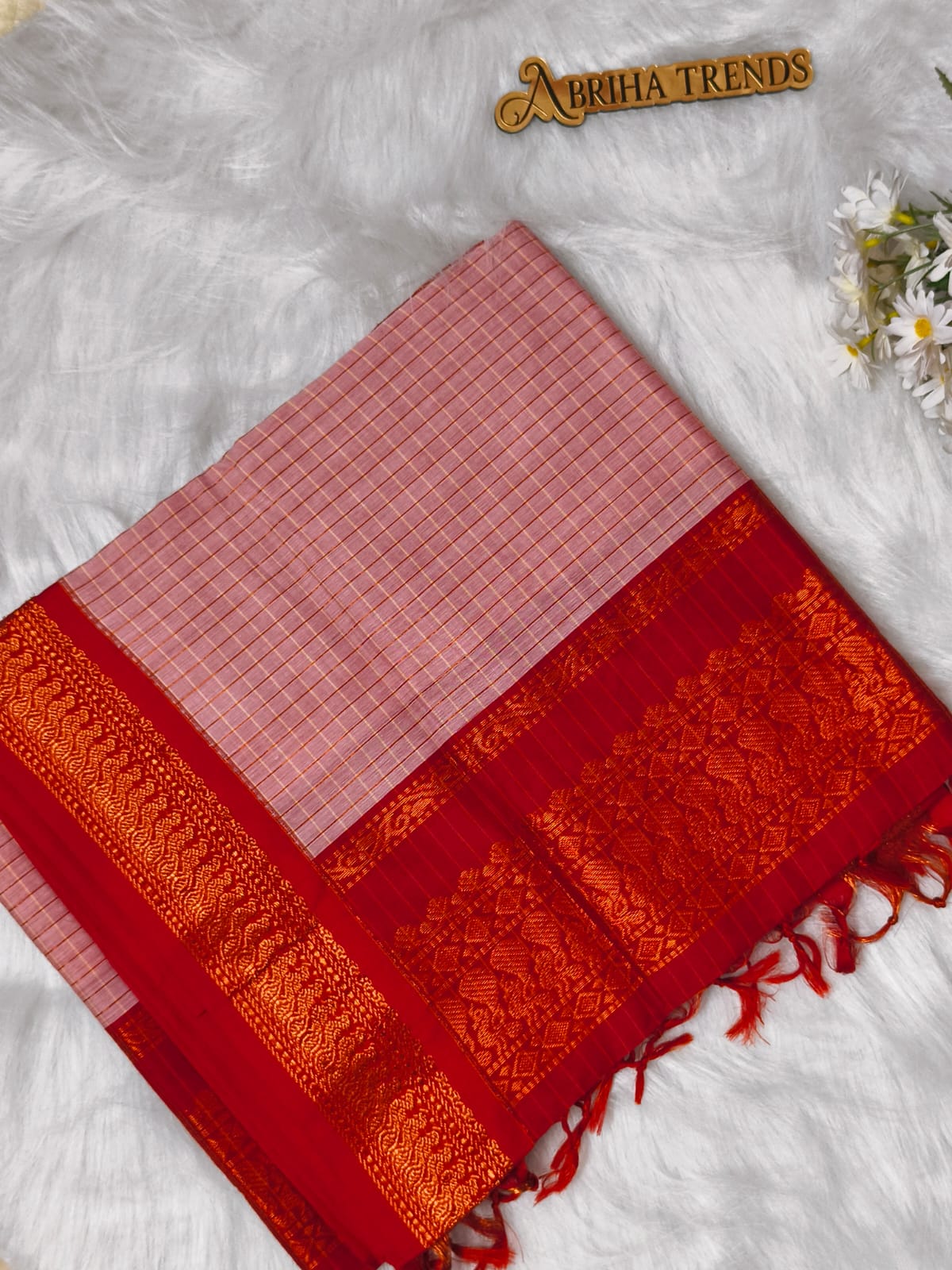A folded red and pink saree with a pink checkered body, a rich red border, and an ornate gold zari brocade, displayed on a white faux fur background with a small gold 'Abhija Trends' logo and white flowers.