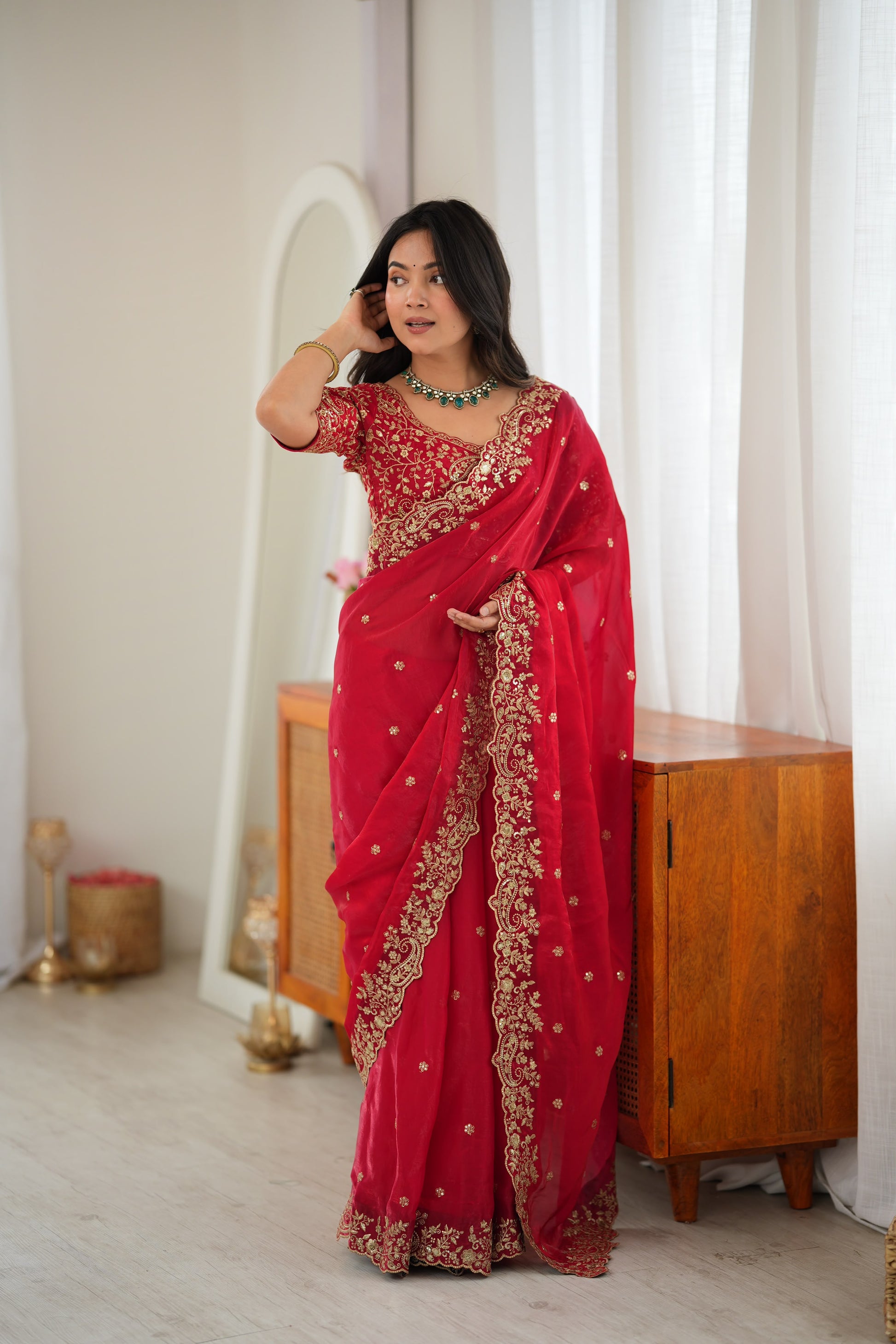 A woman wearing a bright red saree with gold floral embroidery and a matching embroidered blouse, standing in a softly lit indoor room with curtains, a wooden cabinet, and a mirror.