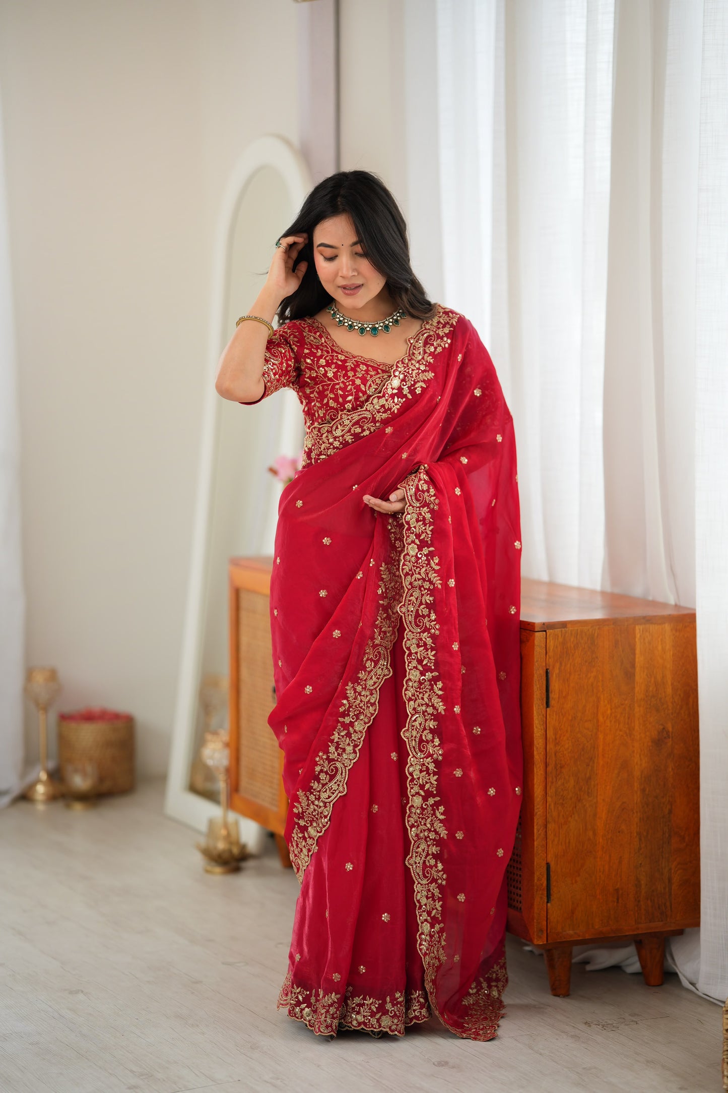 A woman wearing a red saree with gold embroidery and a matching embroidered blouse, standing in a bright indoor room with white curtains, a wooden cabinet, and a mirror.