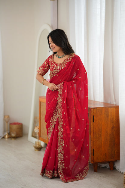 A woman wearing a red saree with gold embroidery and a matching embroidered blouse, standing in a bright indoor room with white curtains and a wooden cabinet.