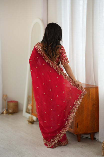 A woman viewed from behind wearing a red traditional sari with gold embroidery and a matching embroidered blouse, standing in a softly lit room beside a wooden cabinet and white curtains.