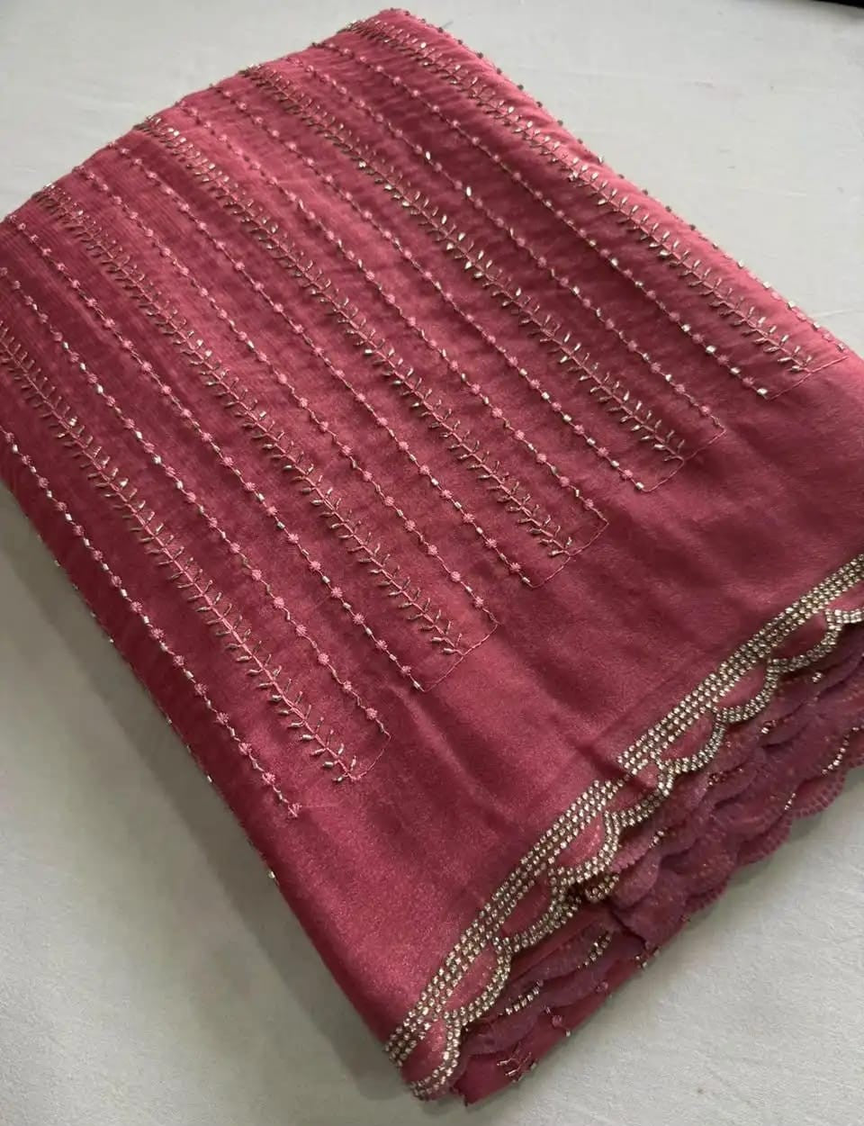 A folded piece of dusty rose pink fabric with vertical metallic gold embroidery and a scalloped beaded border, displayed on a light gray surface.