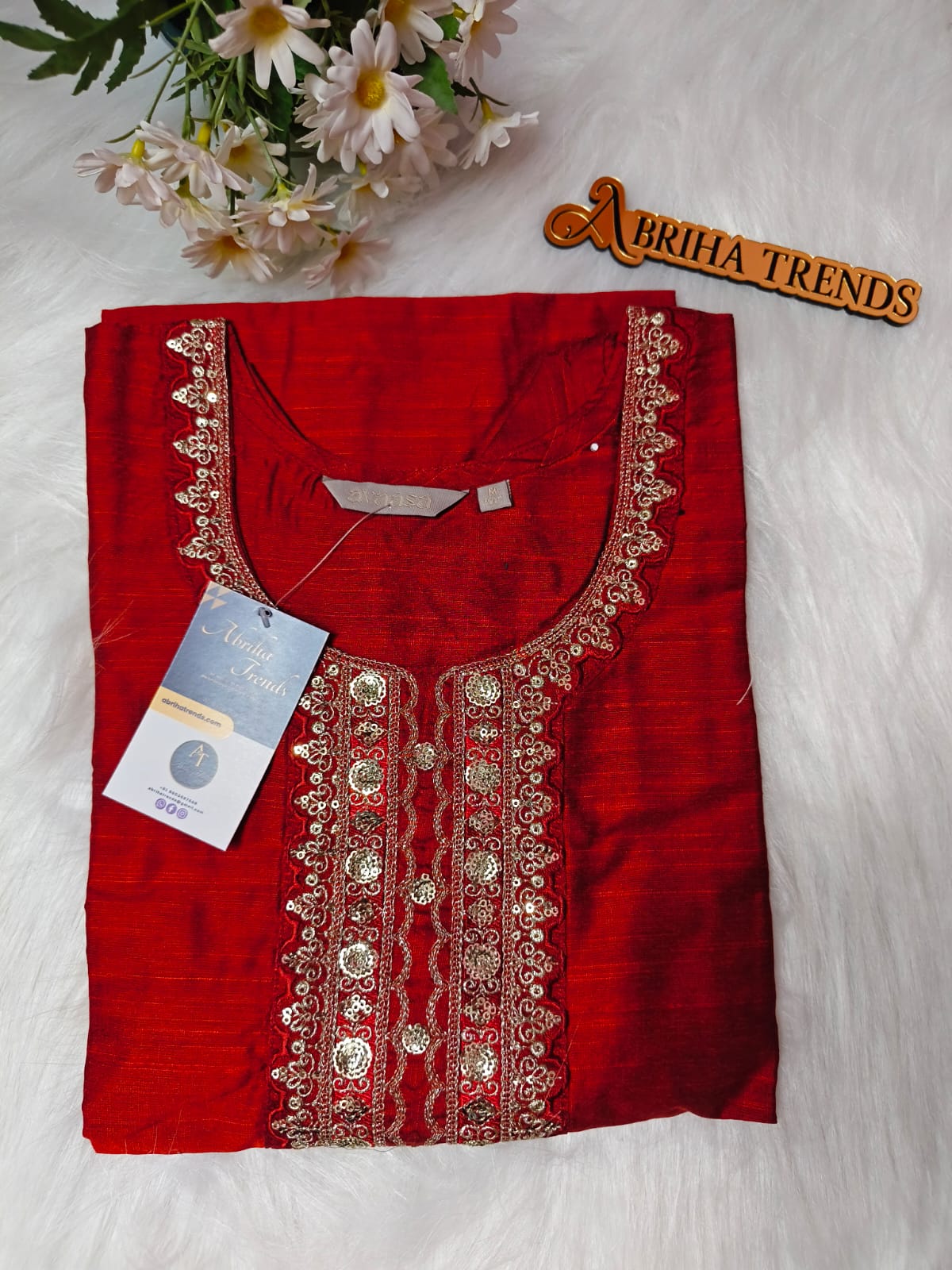 A folded red kurta with ornate gold embroidery and mirror work on the neckline, displayed on a white faux fur background with a floral prop and a 'Abriha Trends' sign.