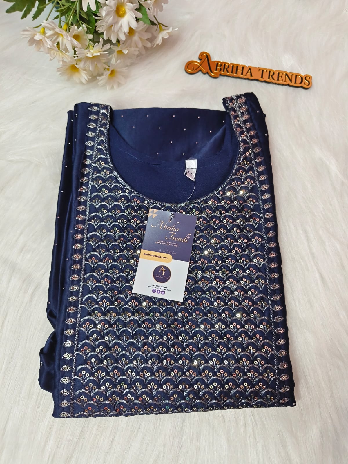 A folded navy blue embroidered kurta set with metallic gold and silver scalloped patterns, displayed on a white faux fur background with a 'Abriha Trends' wooden badge and white flowers.