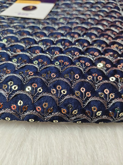 A close-up of a navy blue fabric with an embroidered scallop or fan pattern in metallic silver, decorated with small sequins in gold, pink, and green, laid flat on a white faux fur surface.