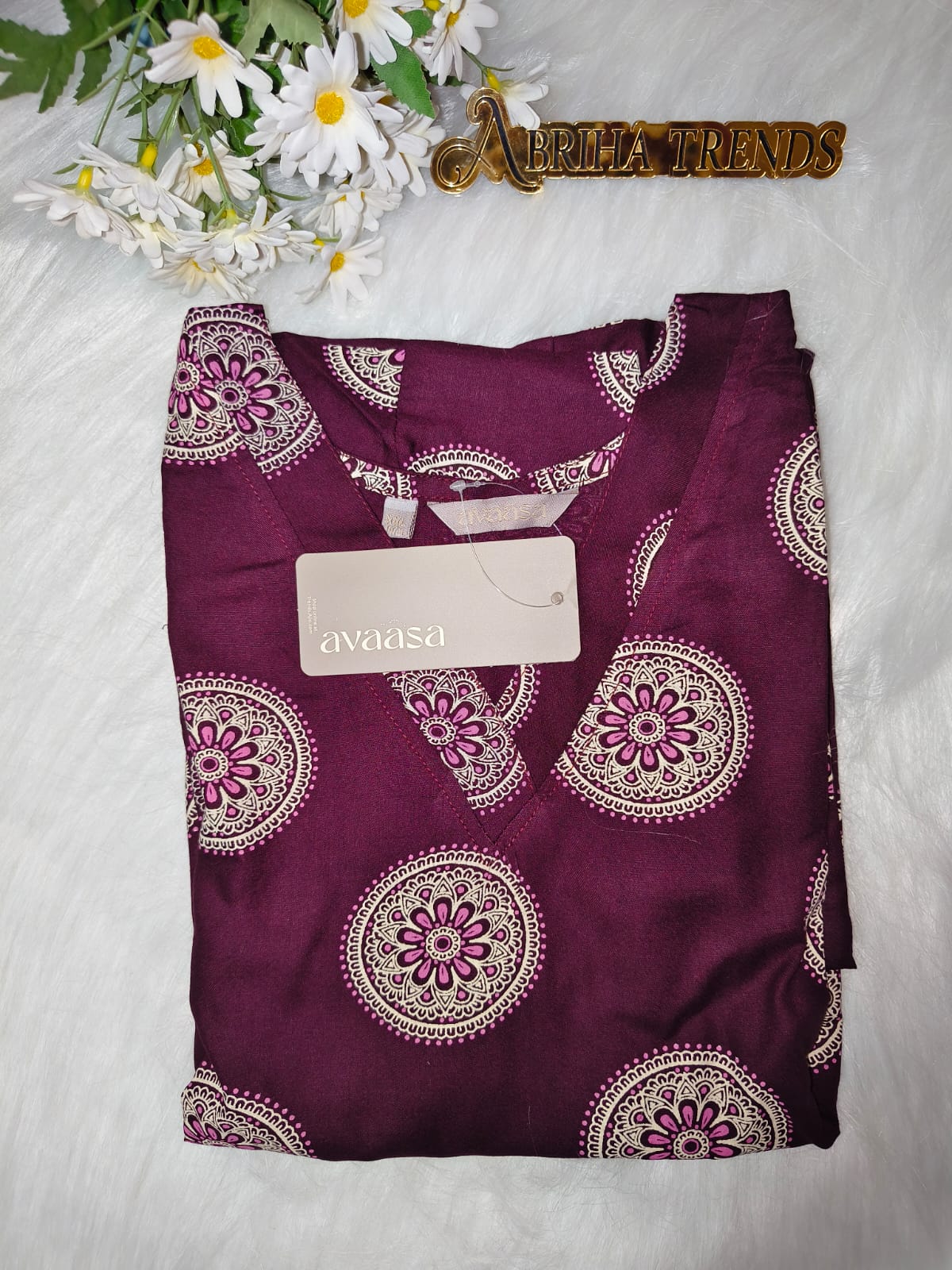 A folded dark purple kurta with a cream and magenta paisley pattern, featuring a matching tie-neck. The item is styled on a white textured background with white daisies and a gold 'Abriha Trends' logo at the top.
