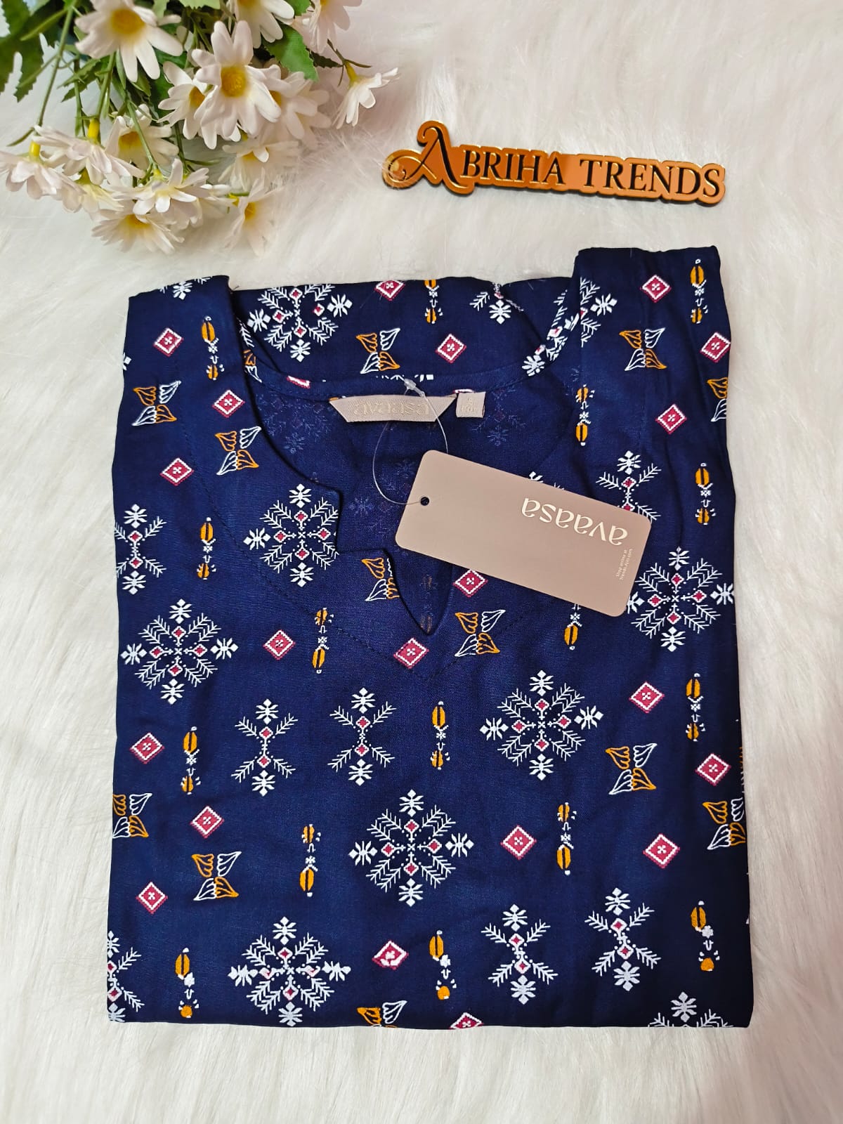 A folded navy blue kurta with a white, pink, and yellow geometric pattern, featuring a beige. The kurta is laid flat on a white faux fur surface next to a wooden 'ABRIHA TRENDS' sign and white daisies.
