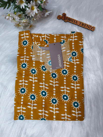 A mustard yellow cotton kurti with a white and teal floral print, folded neatly on a white faux fur surface with a brand tag attached. A wooden brand tag reading 'ABRIHA TRENDS' is visible, along with some white daisies in the top left corner.