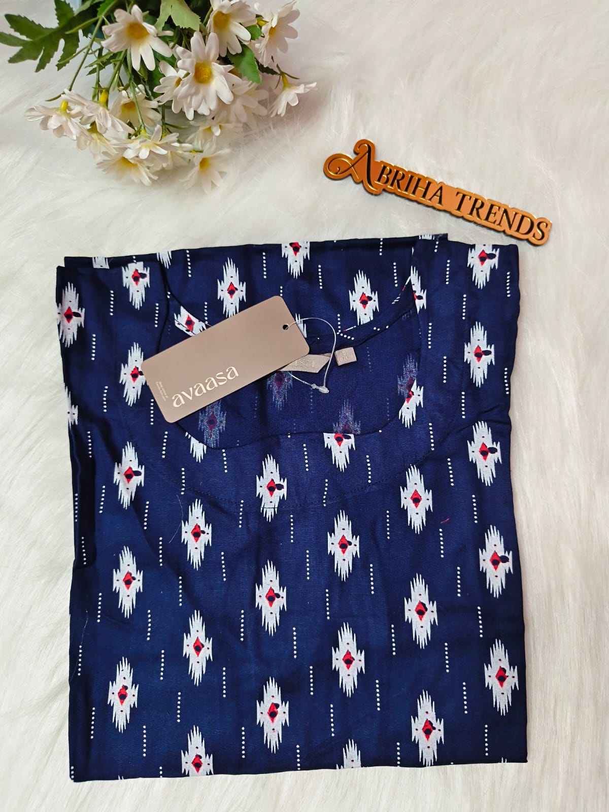 A folded navy blue kurta with a white and red ikat pattern and a decorative wooden 'ABRIHA TRENDS' sign on a white faux fur background with white daisies.