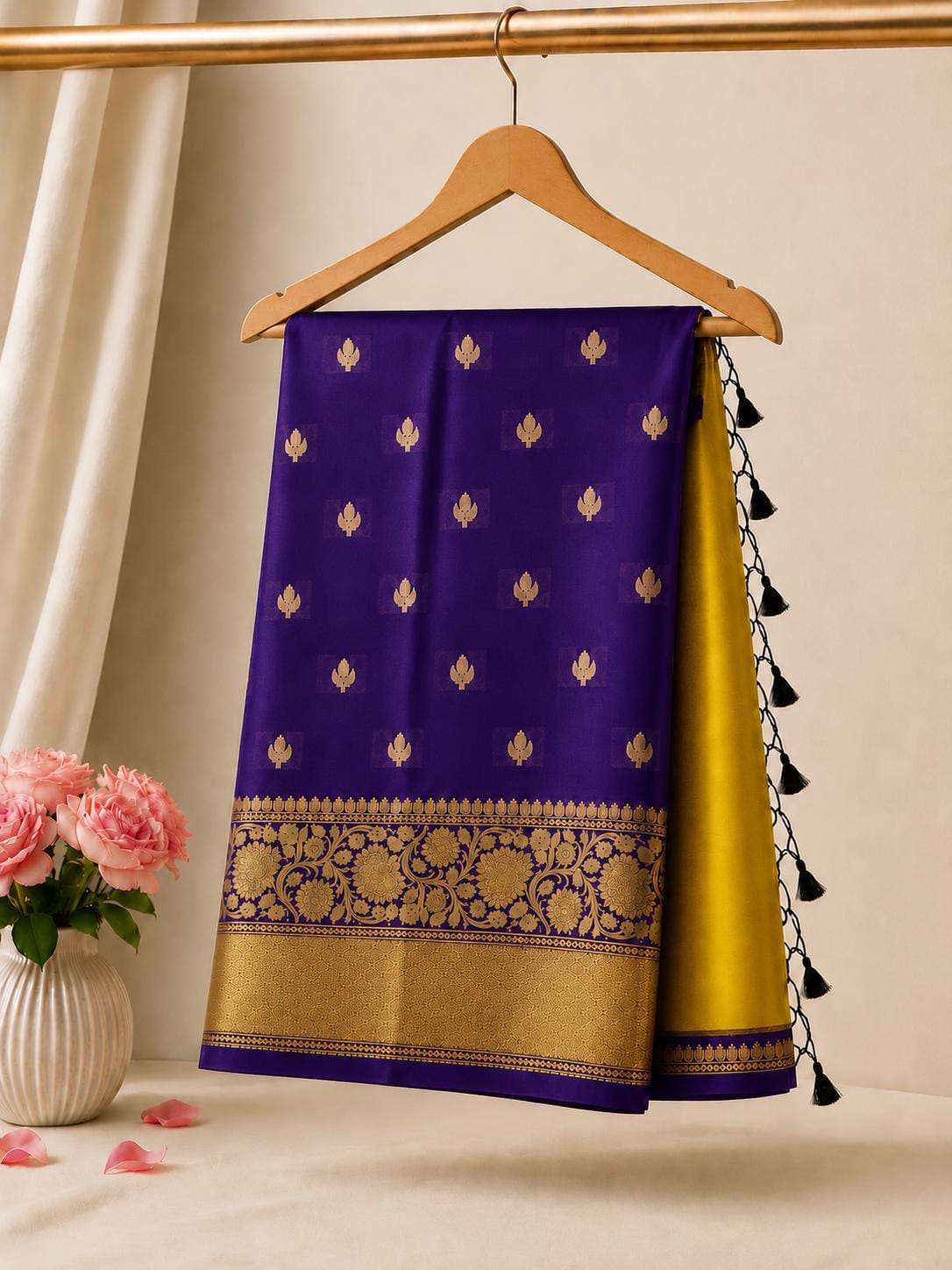 A purple and gold Banarasi silk saree draped elegantly on a wooden hanger, with a gold side panel showing black tassel ties, set against a warm beige background with a vase of pink roses in the corner.