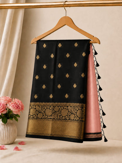 A black and pink saree draped elegantly on a wooden hanger, featuring gold zari motifs, a detailed gold border, and black tassel ties, set against a warm beige background with a vase of pink roses.