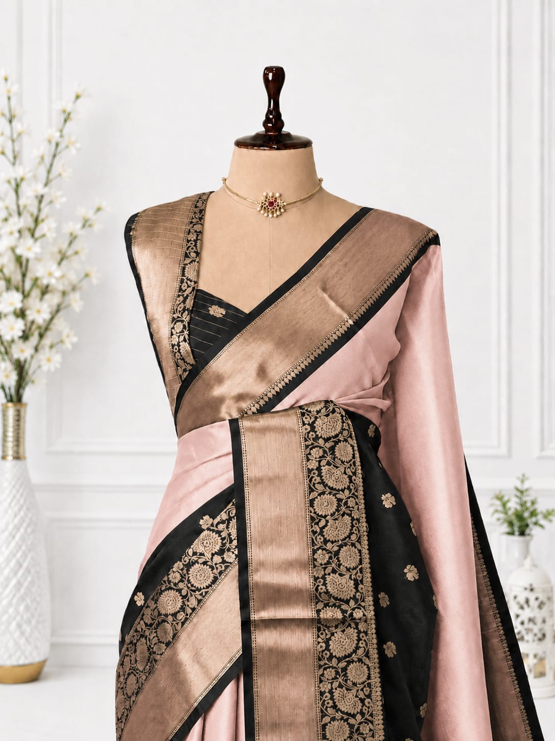 A pink and black silk saree with intricate gold zari embroidery, displayed on a mannequin torso with a matching blouse. The background is a clean white paneled wall with decorative floral vases.