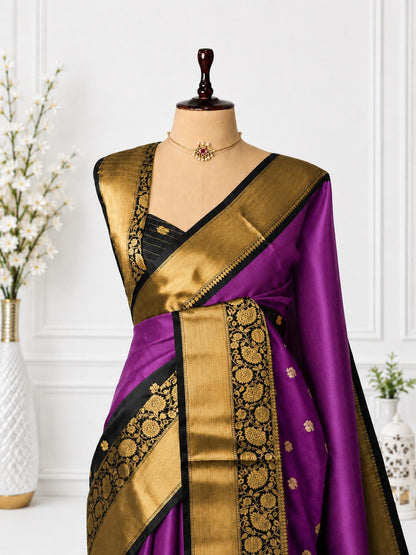 A purple silk saree with a wide gold and black zari border, draped on a mannequin torso. The blouse is black with gold embroidery, and the setting includes a decorative white wall, a vase of white flowers, and a small potted plant.