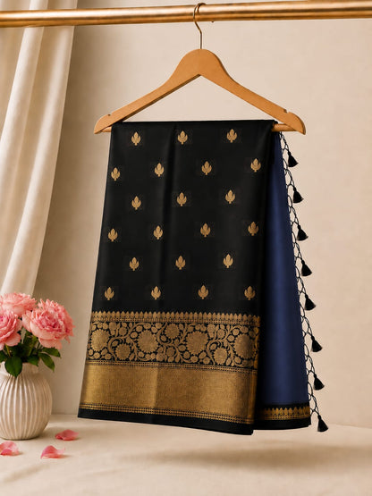 A black and navy blue silk saree draped on a wooden hanger, featuring gold zari embroidery, a decorative border, and black tassels, set against a warm beige background with a vase of pink roses.