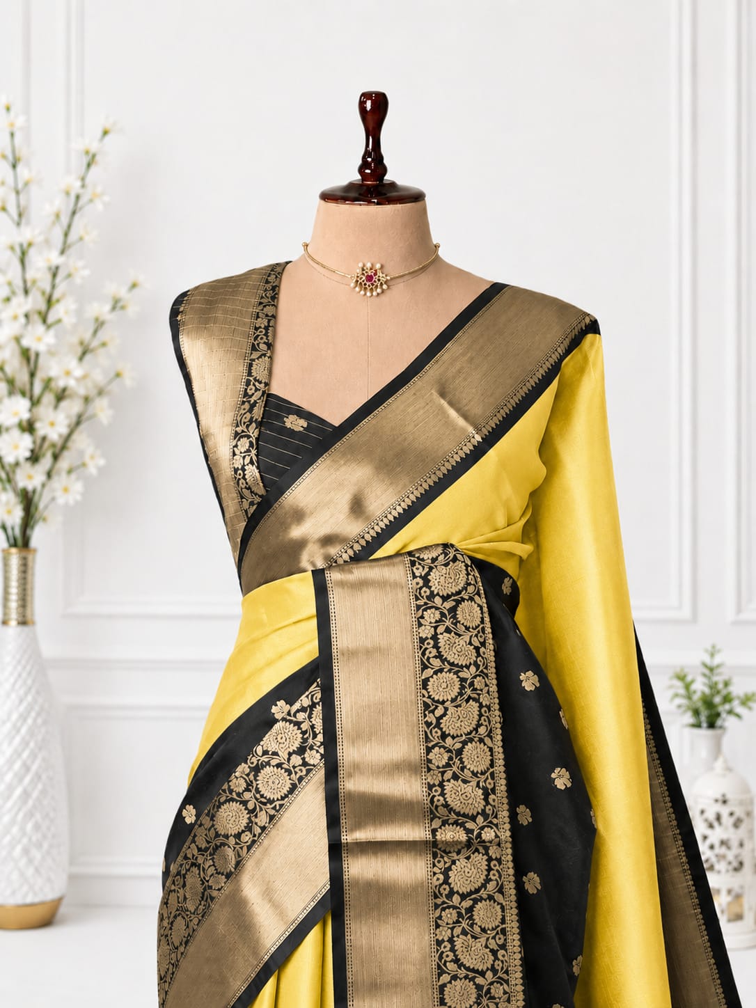 A yellow and black silk saree with intricate gold zari brocade patterns, displayed on a mannequin torso with a matching black blouse and necklace.