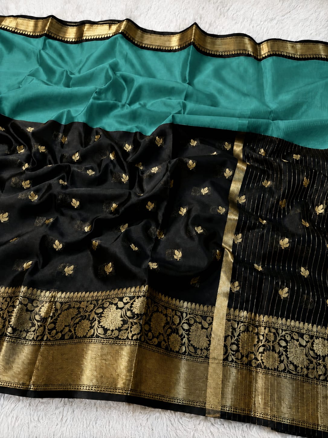 Black and teal green silk saree with intricate gold zari brocade borders and small gold floral motifs, laid flat on a white textured surface.