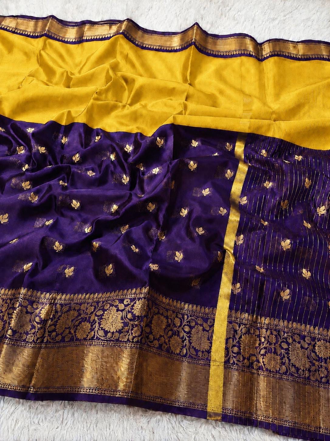 A purple and gold Banarasi silk saree with a yellow-gold pallu, gold zari motifs, and a wide gold border, laid flat on a white textured surface.
