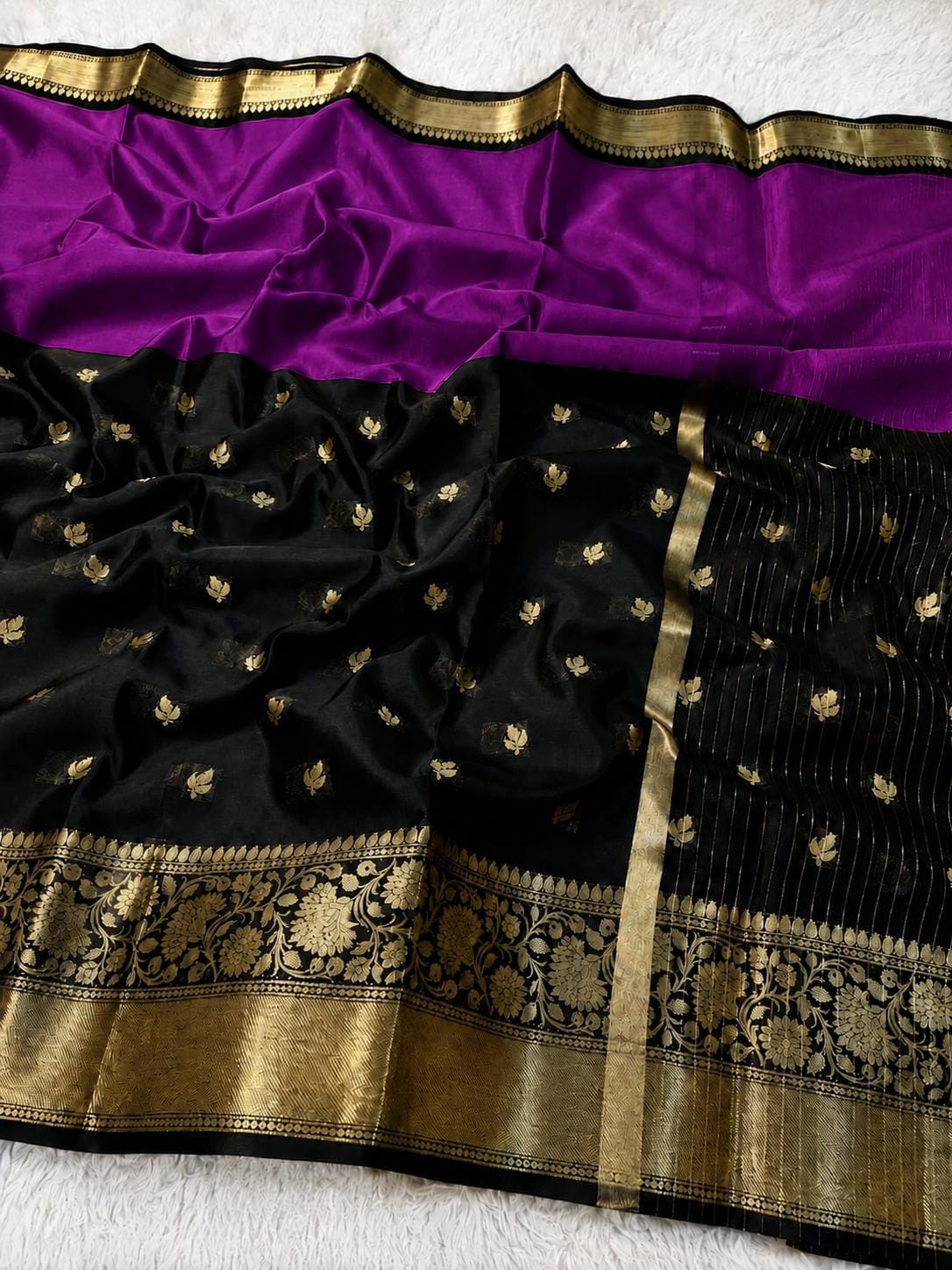 A black silk saree with a vibrant purple border and a wide gold zari brocade pallu, laid flat on a white textured surface.