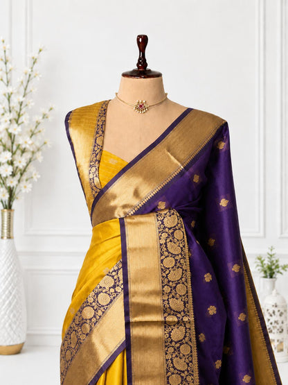 A purple and yellow silk saree with intricate gold zari embroidery, displayed on a mannequin torso with a matching blouse. The background is a clean white paneled wall with decorative floral vases.