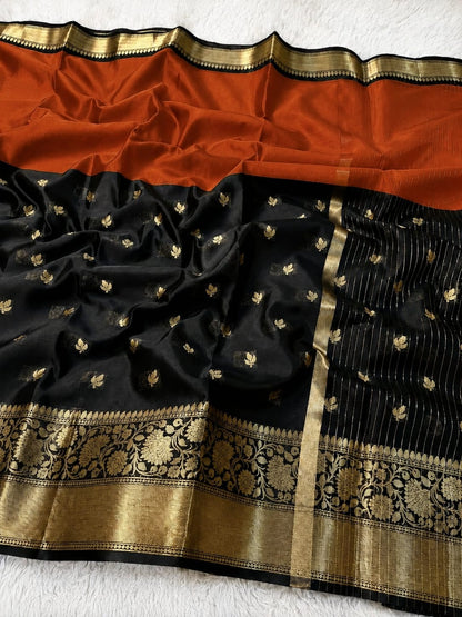 A black silk saree with a rust orange border, decorated with small gold floral motifs and featuring an ornate gold zari brocade pallu and border.