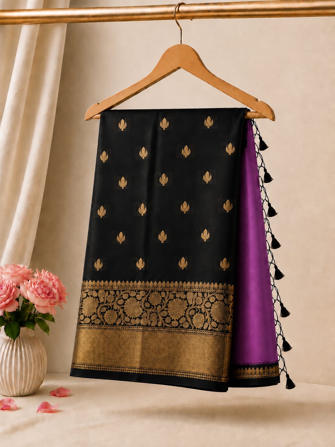 A black and purple silk saree with gold zari embroidery hanging on a wooden hanger, styled with pink roses in a vase against a neutral beige background.