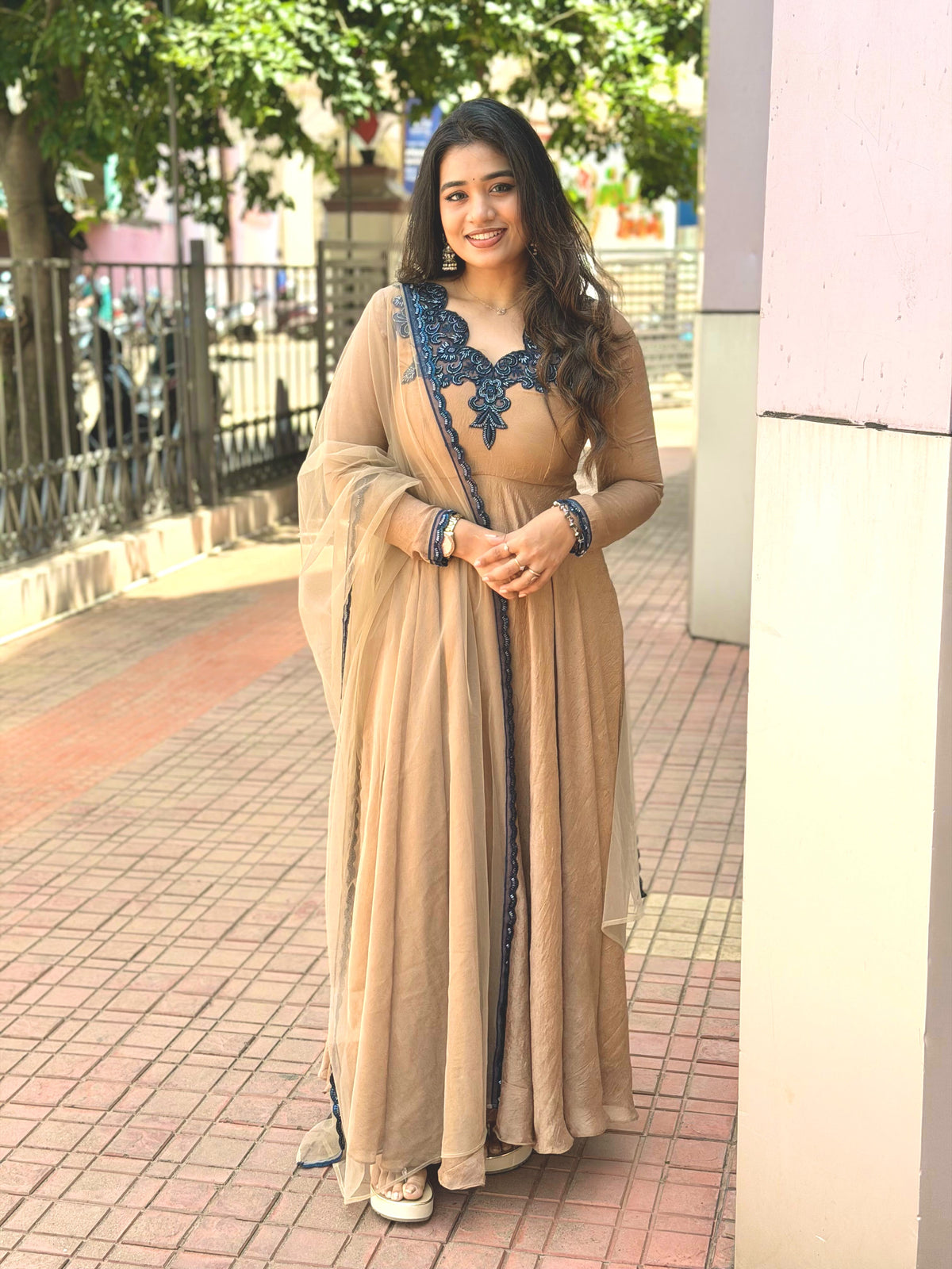 A woman wearing a beige floor-length traditional dress with dark blue embroidery and a matching sheer dupatta, standing on a paved outdoor walkway.