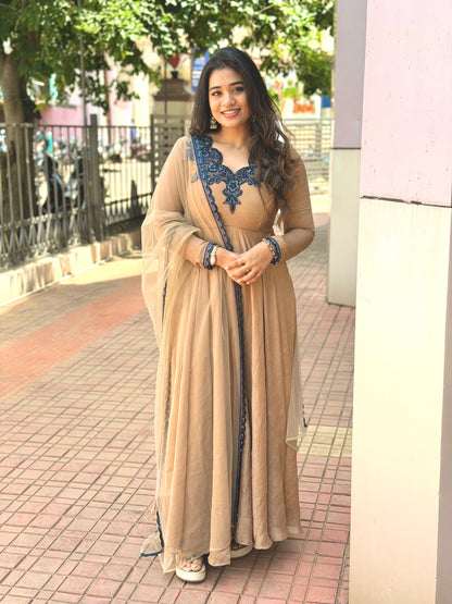 A woman wearing a beige floor-length traditional dress with dark blue embroidery and a matching sheer dupatta, standing on a paved outdoor walkway.