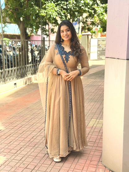 A woman wearing a beige traditional Anarkali gown with blue embroidery and a matching sheer dupatta, standing outdoors on a paved walkway.