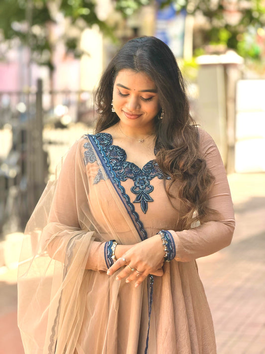 A woman wearing a beige traditional outfit with dark blue embroidered lace details on the bodice and a matching sheer dupatta, standing outdoors in a softly blurred street setting.