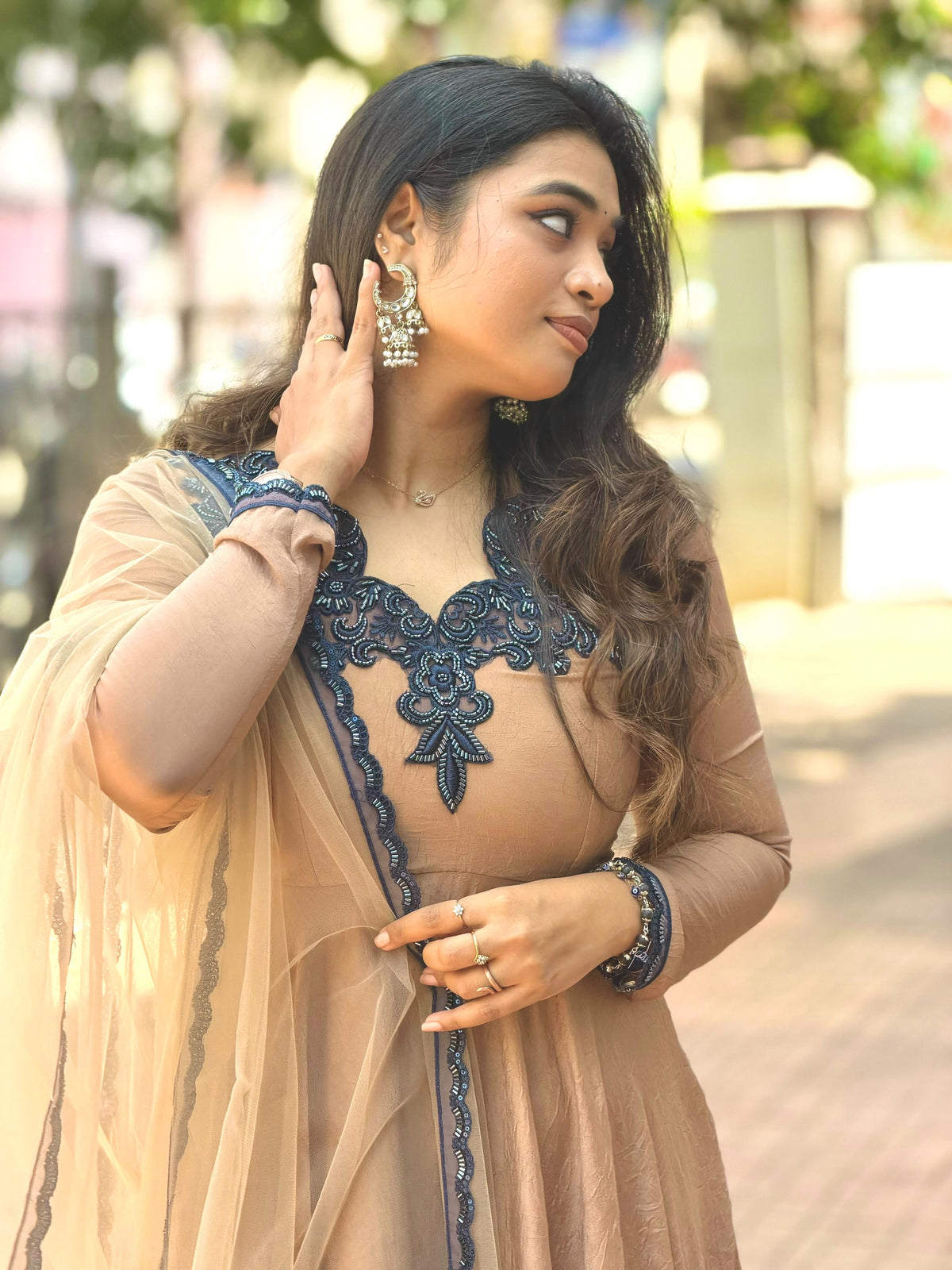 A woman wearing a beige traditional outfit with a dark blue embroidered neckline and matching dupatta, photographed outdoors with a blurred background.