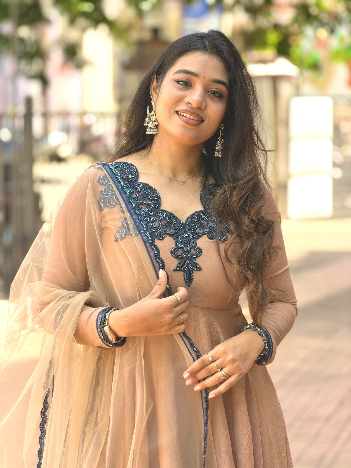 A woman wearing a peach-beige traditional dress with dark navy blue embroidery and a matching sheer dupatta, standing outdoors in a blurred street setting.