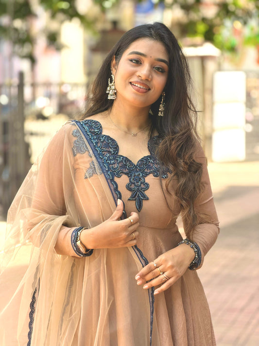 A woman wearing a peach-beige traditional dress with dark navy blue embroidery and a matching sheer dupatta, standing outdoors in a blurred street setting.