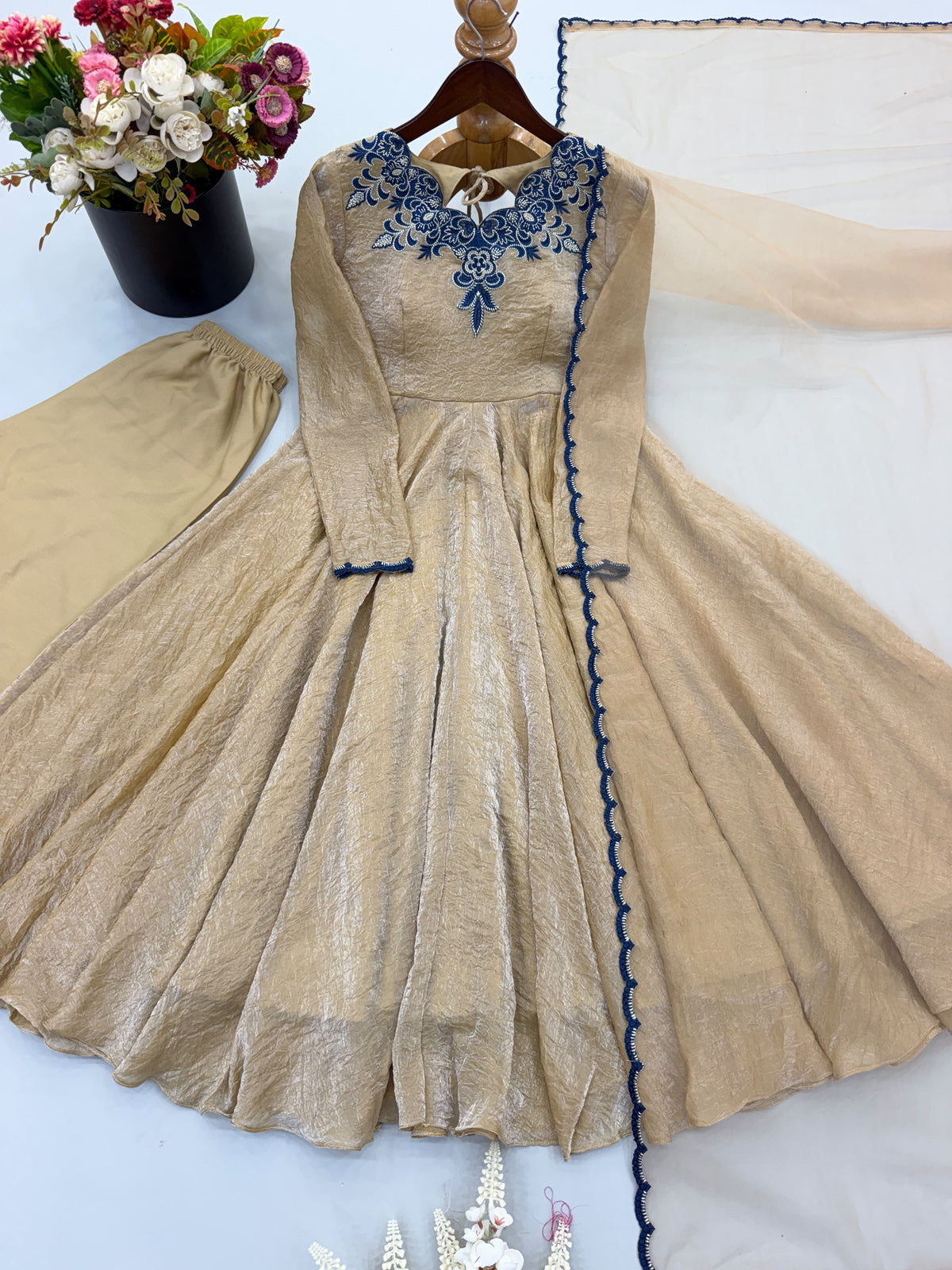 A beige long-sleeve Anarkali-style dress with blue embroidered neckline and blue trim details, displayed on a hanger with matching beige pants and a sheer beige fabric draped beside it on a white surface.