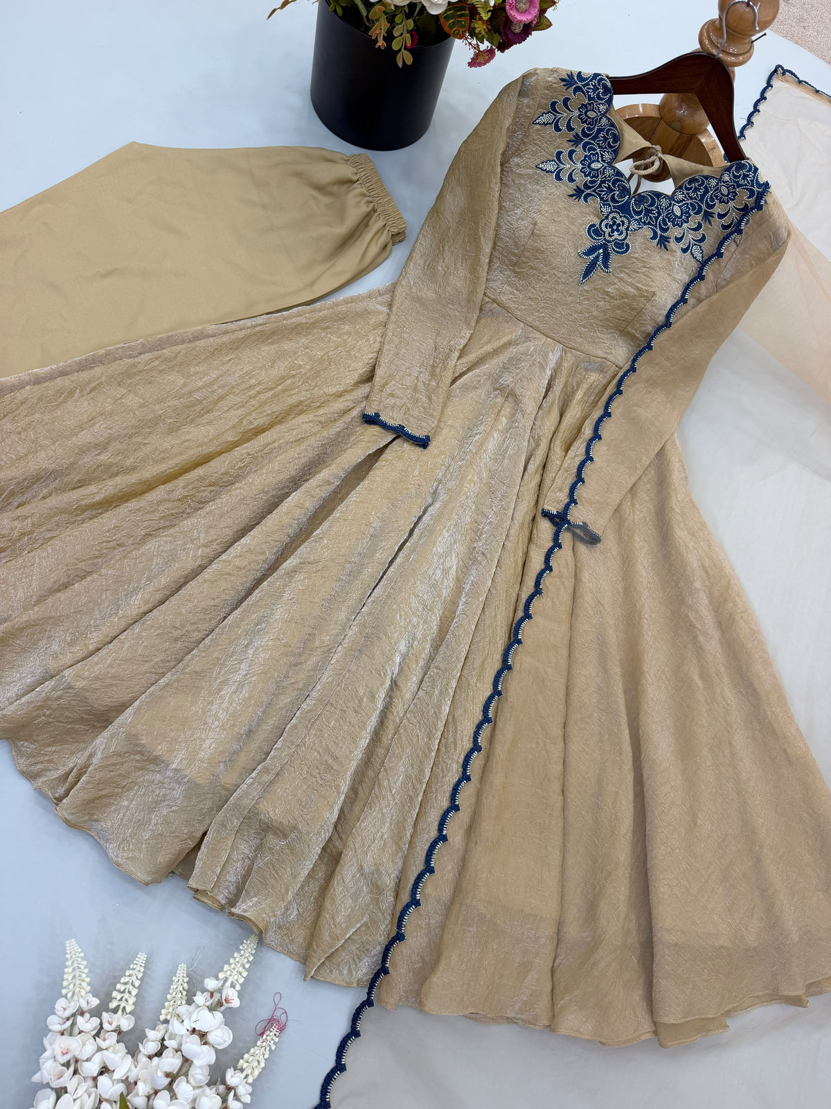 A beige long-sleeve dress with blue floral embroidery around the neckline and a blue scalloped trim along the front, displayed on a hanger with a matching beige fabric piece and decorative flowers in the background.
