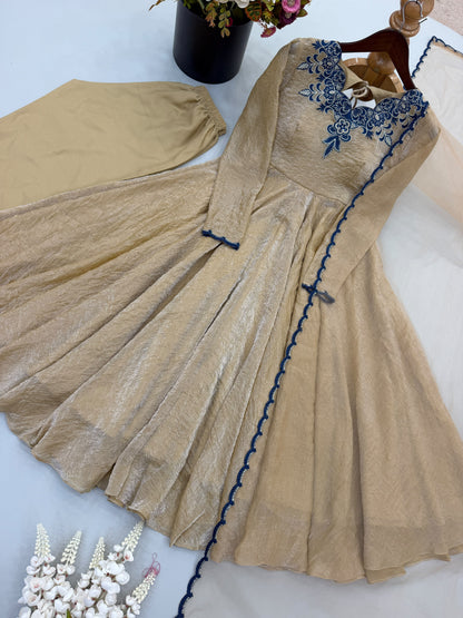 A beige long-sleeve dress with blue floral embroidery around the neckline and a blue scalloped trim along the front, displayed on a hanger with a matching beige fabric piece and decorative flowers in the background.