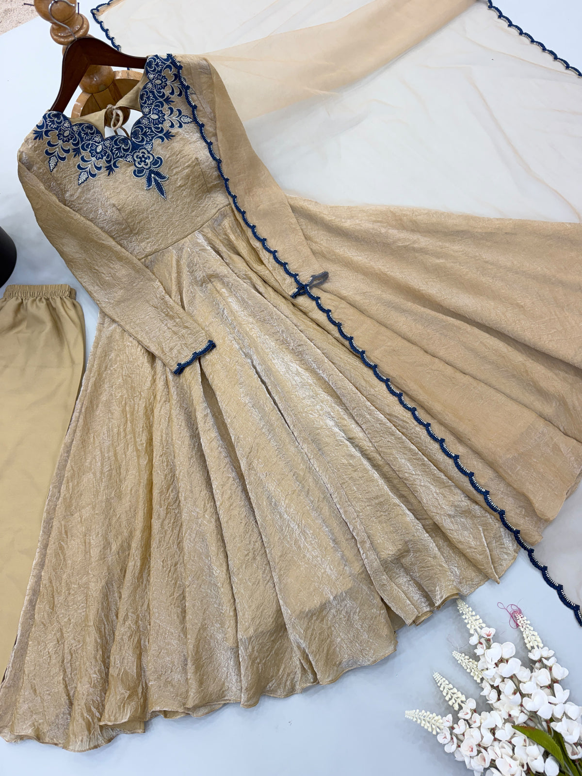A beige long-sleeve Anarkali-style dress with blue floral embroidery at the neckline and blue scalloped trim, shown laid flat with matching beige pants and a sheer dupatta on a white background.
