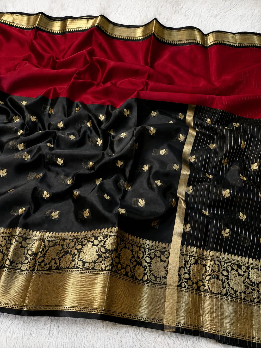 A black silk saree with a rich red pallu, adorned with small gold floral motifs and a wide ornate gold zari border, laid flat on a white textured background.
